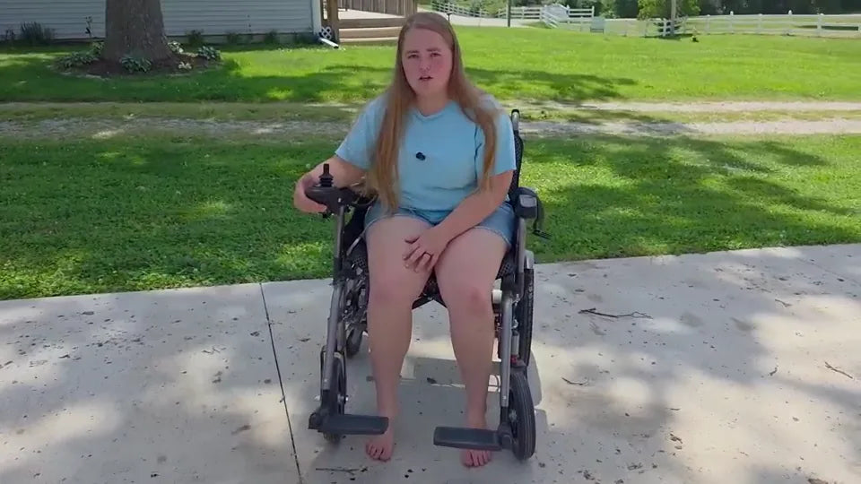 Real YS16-1 MAX rider seated in a stationary chair outdoors with both feet on the ground and the foot supports positioned to either side of the feet.